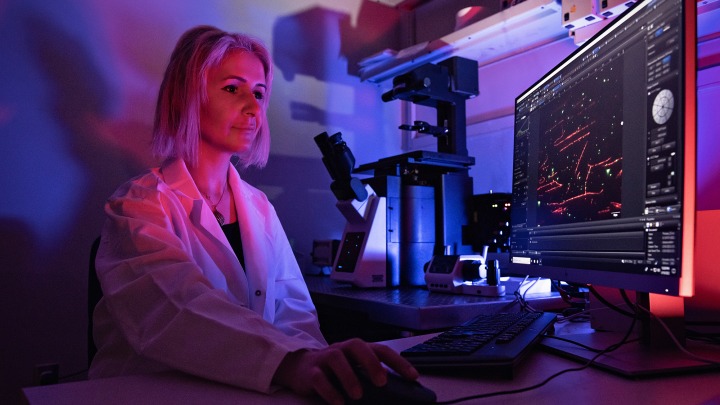 A person in a lab coat works at a computer in a dimly lit lab with a large microscope nearby.